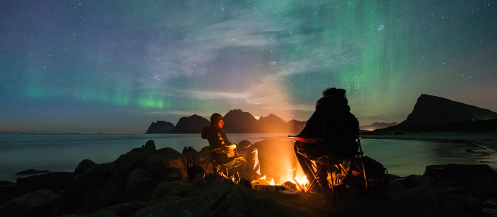 A couple enjoy a campfire while the Northern Lights dance in the sky above