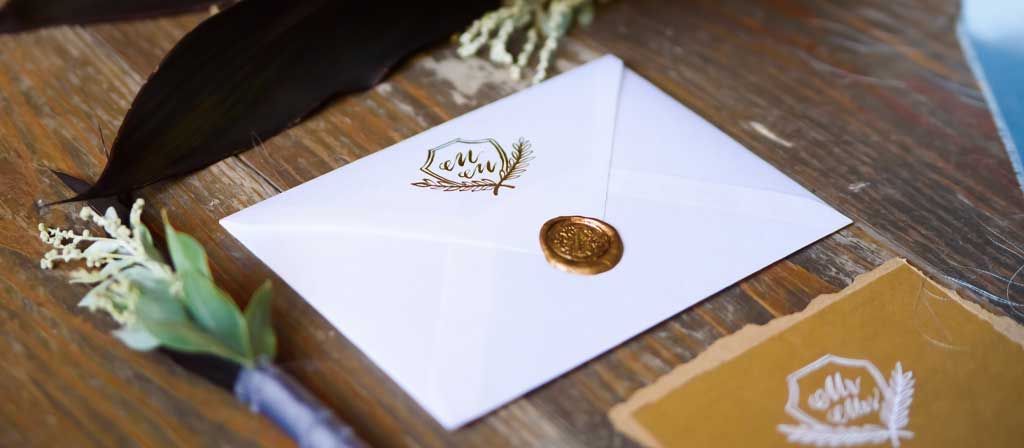 Un sobre con un sello decorativo que contiene una invitación yace sobre una superficie de madera con arreglos florales