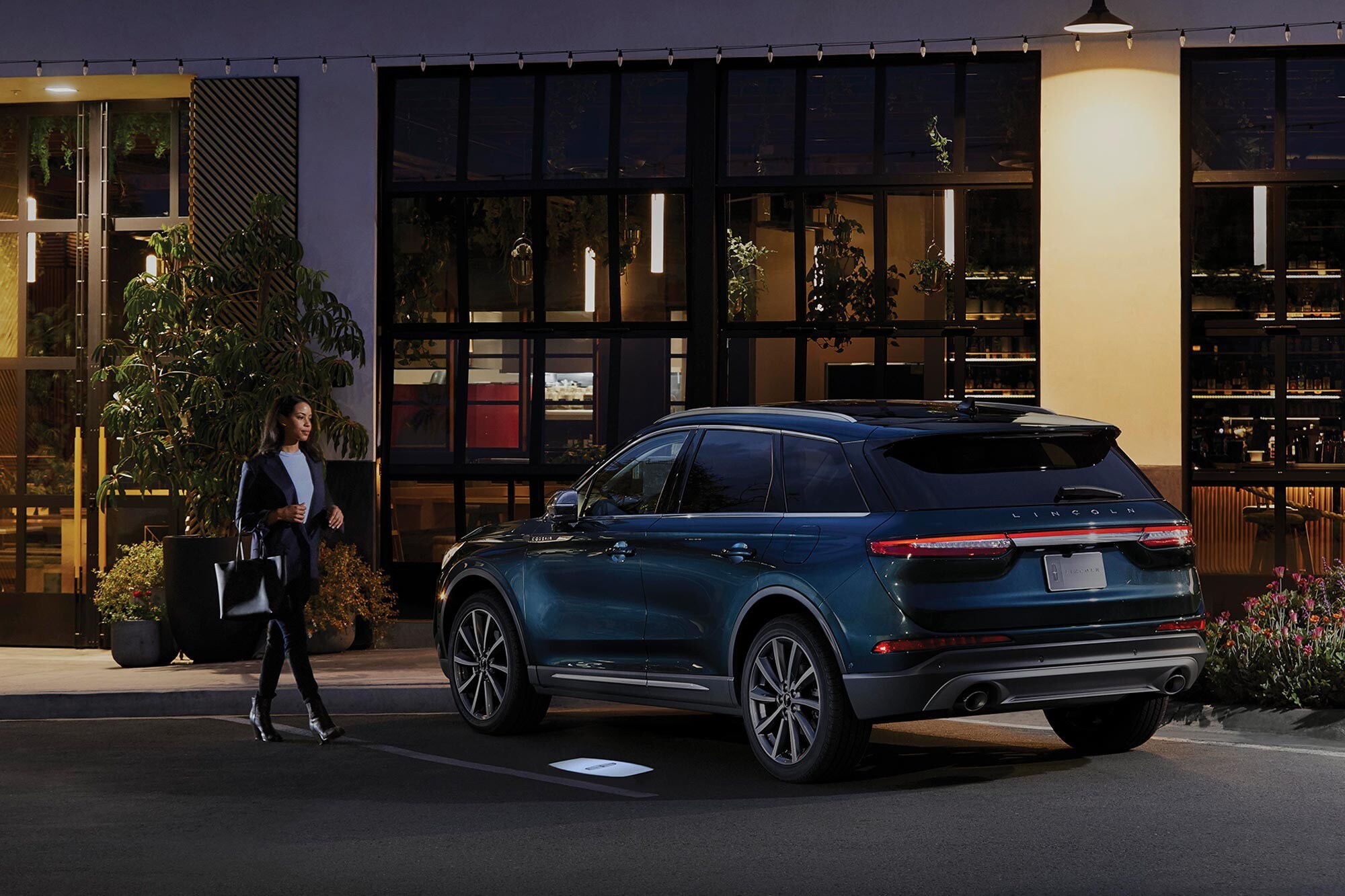A woman walking towards a blue Lincoln Corsair at night time with a lit up Lincoln logo displaying on the pavement.