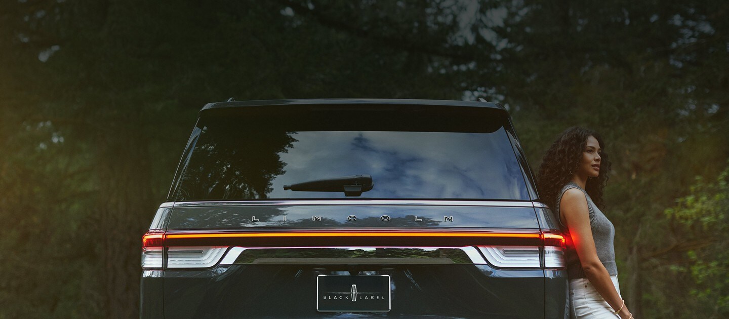 A woman is leaning against a 2023 Lincoln Navigator® Black Label SUV as horizontal rear taillamps light up with a warm glow.