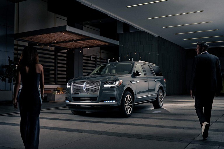 A man and woman in formal wear are approaching a 2023 Lincoln Navigator® SUV at night as it lights with the Lincoln Embrace.
