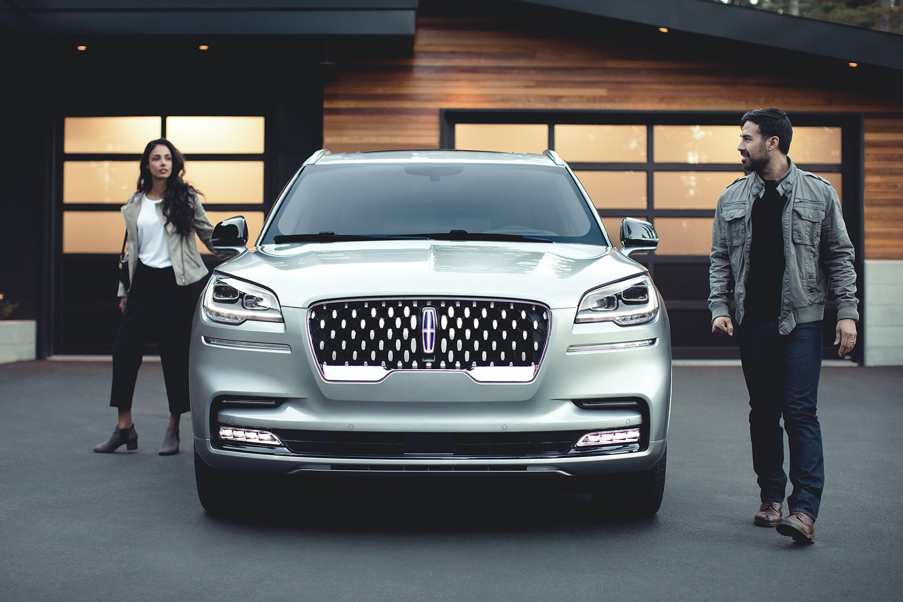 A couple walking around opposite sides of a Lincoln vehicle parked in the driveway of a modern home.