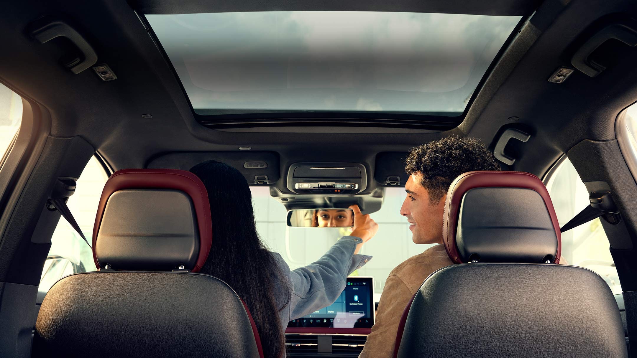 A man and woman sitting in their vehicle.  The woman is adjusting the rear-view mirror.