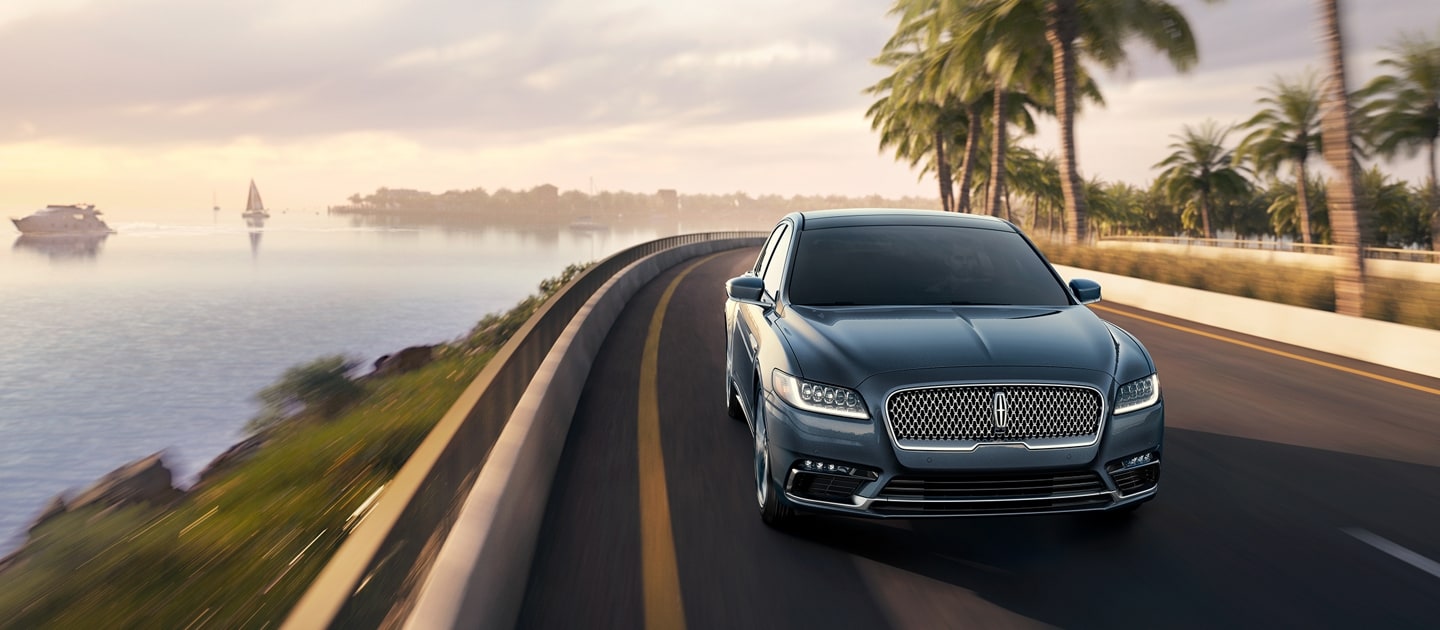 A Lincoln Continental is being driven along a seaside coast with a yacht and sailboat in the water and palm trees along the pavement