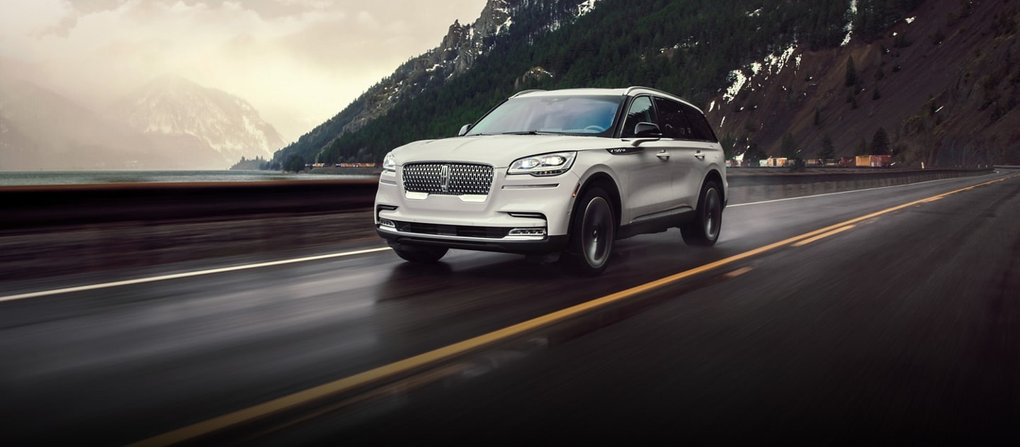 The Lincoln Aviator driving down the road past a mountain range and some water.