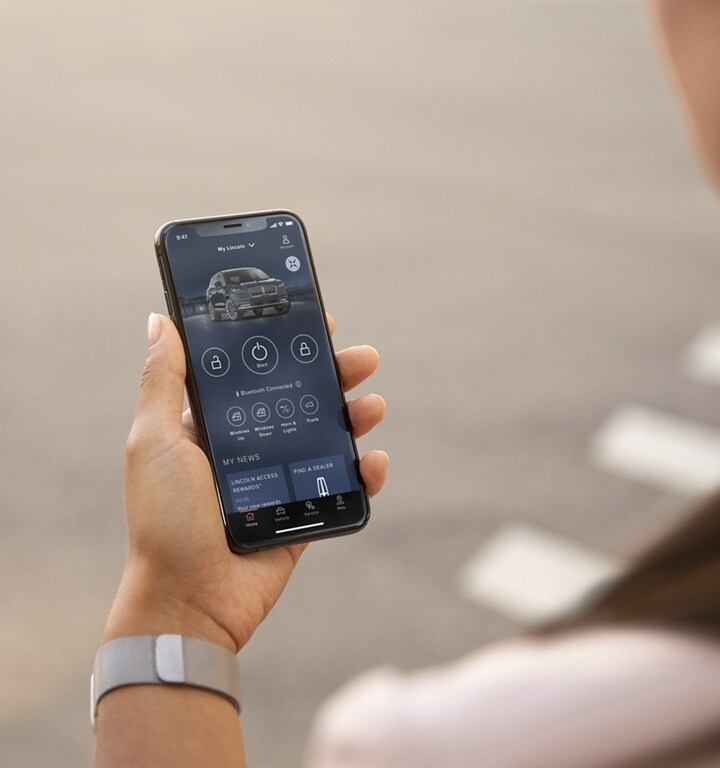 A woman is standing at a sunny window interacting with the Lincoln Way™ App on her smartphone device