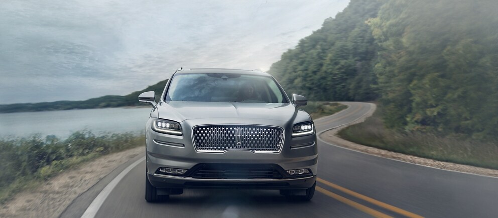 The front end of the 2023 Lincoln Nautilus® SUV is shown being driven around a curve on a lakeside road