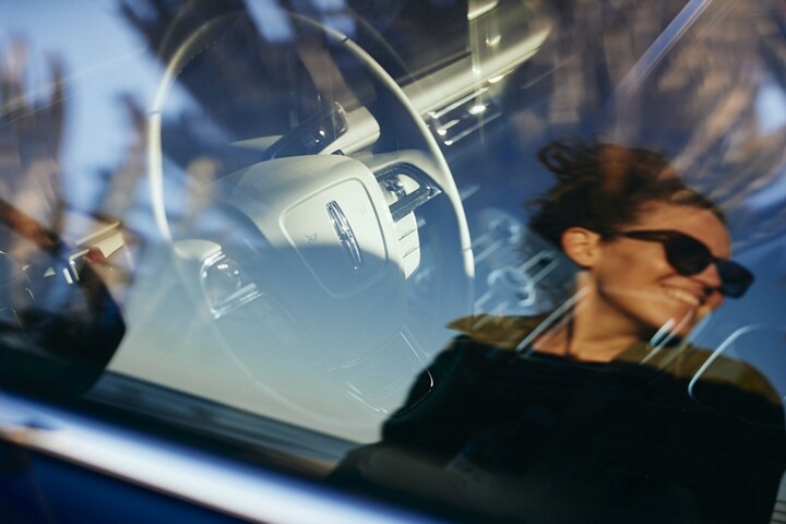 The steering wheel of the Lincoln Nautilus is shown here through the driver’s window with the reflection of a woman.