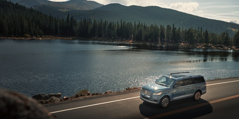 A blue Lincoln Navigator driving in the foreground by a serene lake with mountains behind.