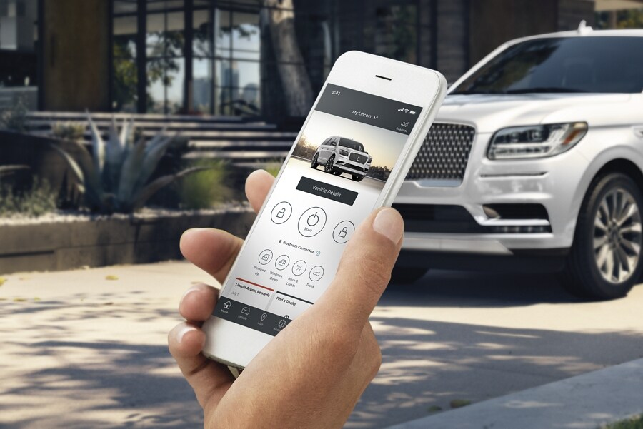 A hand is holding a smartphone with the Lincoln Way App displayed on the screen in front of a 2021 Lincoln Navigator parked in the driveway of a home