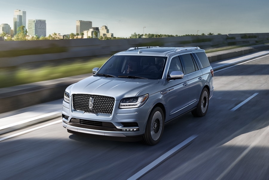 A 2021 Lincoln Black Label Navigator in Chroma Crystal Blue is being driven on a freeway near sunlit fields and a city in the background
