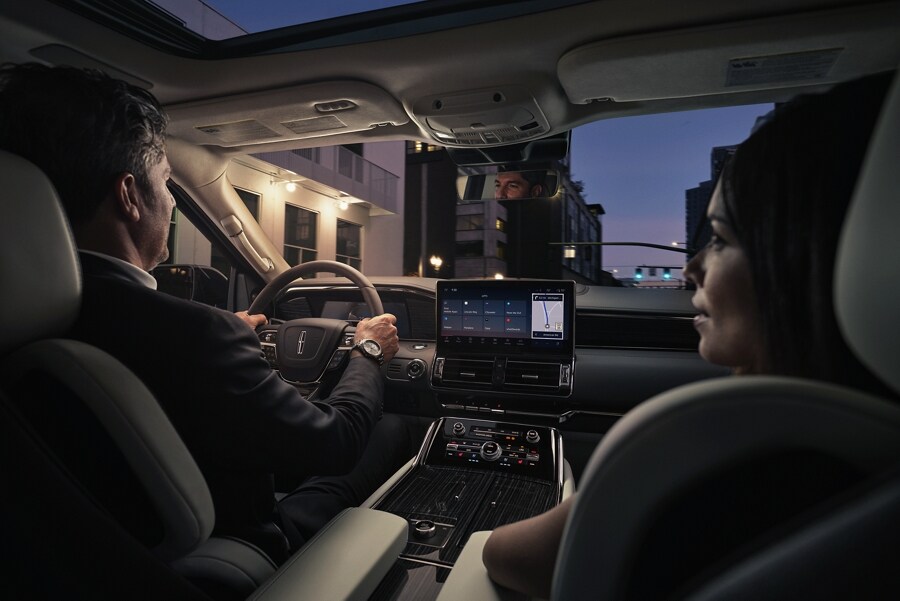 A man and woman are relaxing inside a 2023 Lincoln Navigator® SUV as they drive through a downtown street at night.