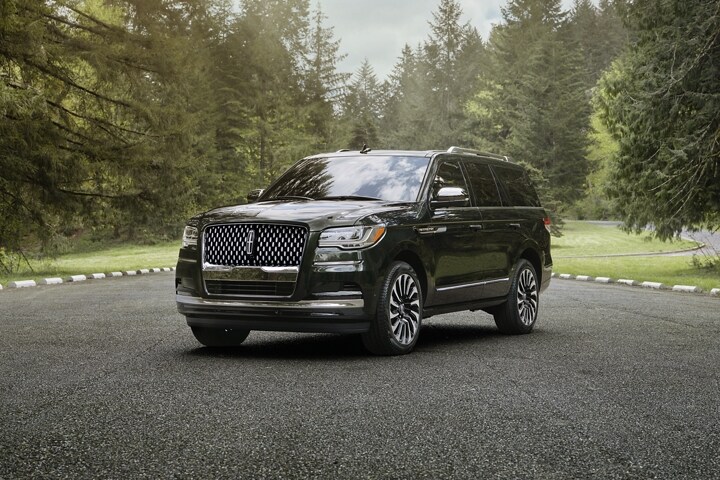 A 2023 Lincoln Black Label Navigator® SUV in Manhattan Green is parked on pavement near a scenic forest full of pine trees.