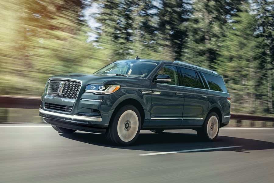 A 2023 Lincoln Navigator® SUV is being driven on a highway lined with tall pine trees as it bathes in sunlight.