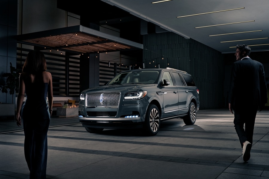 Un hombre y una mujer con ropa formal se acercan a una SUV Lincoln Navigator® 2023 por la noche mientras se enciende con Lincoln Embrace.