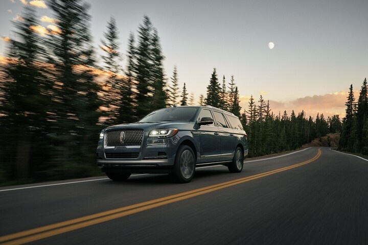 A 2023 Lincoln Navigator® SUV is being driven along a road lined with tall pine trees with a faint moon in the sky at dusk.
