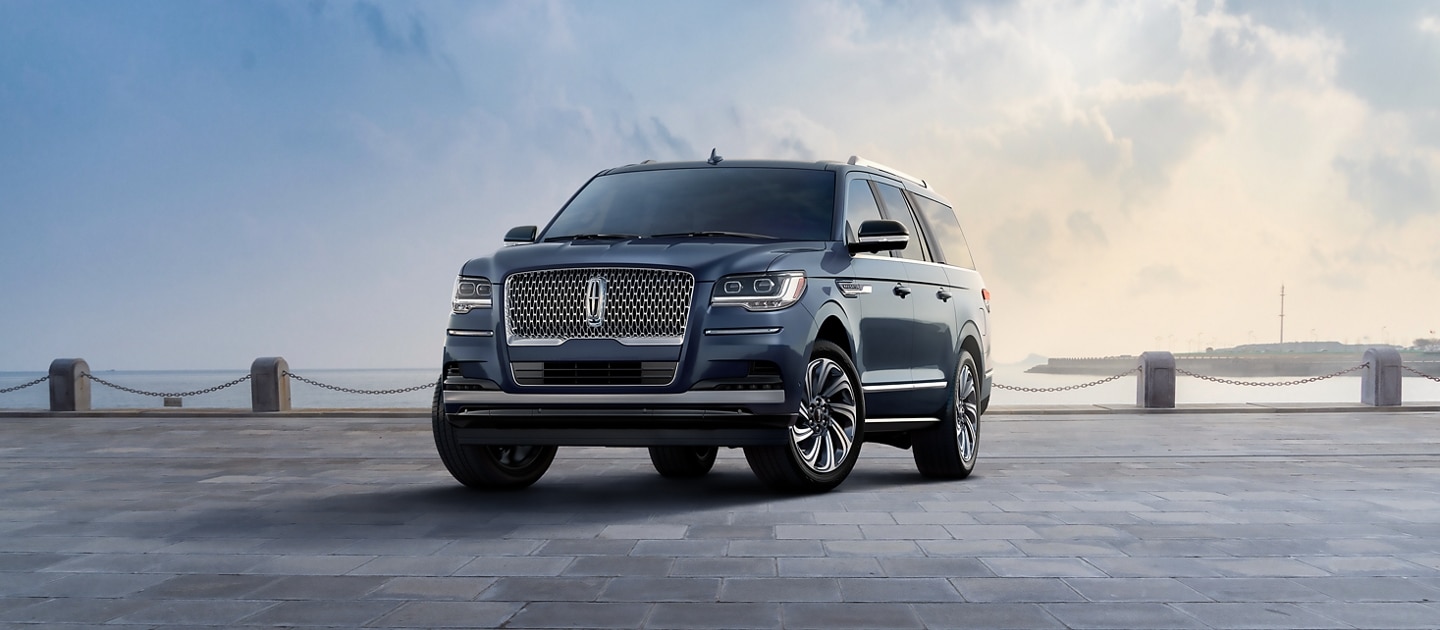 A 2023 Lincoln Navigator® Reserve L model SUV in Ocean Drive Blue is parked near a bay beneath a cloudy sky.