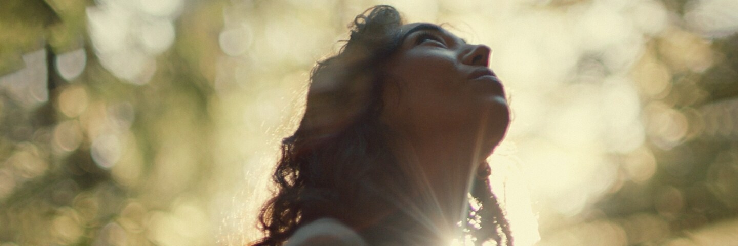 Una mujer contemplando el cielo en un bosque frondoso, mientras la luz del sol se proyecta entre los árboles.