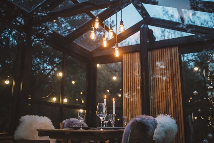 Una mesa al aire libre está armada con vasos, velas y mantas que brillan con bombillas Edison en el techo al atardecer.