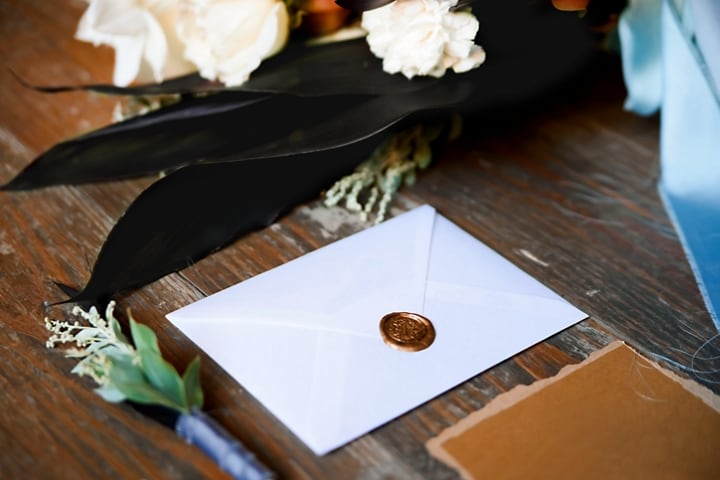 Un sobre blanco con sello de cera dorado sobre una mesa de madera con flores.