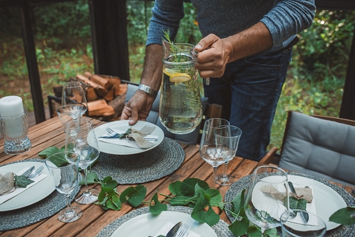 An outdoor table is set with plates and vine garland as a person brings a pitcher of lemon water to set on the table.