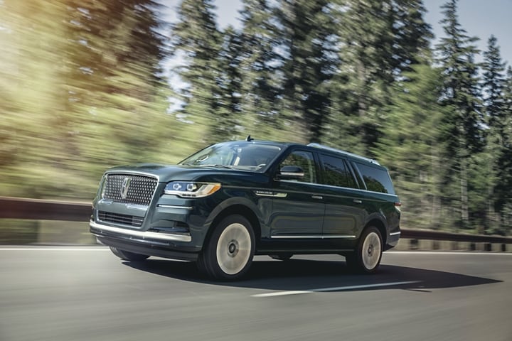A 2024 Lincoln Navigator® SUV in Flight Blue is being driven around a curve surrounded by greenery.