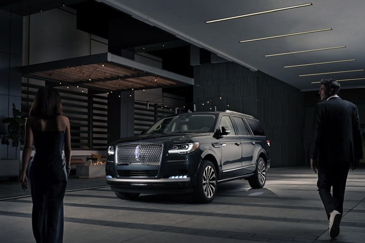Un hombre y una mujer con ropa formal se acercan a una SUV Lincoln Navigator® 2024 por la noche mientras se enciende con Lincoln Embrace.