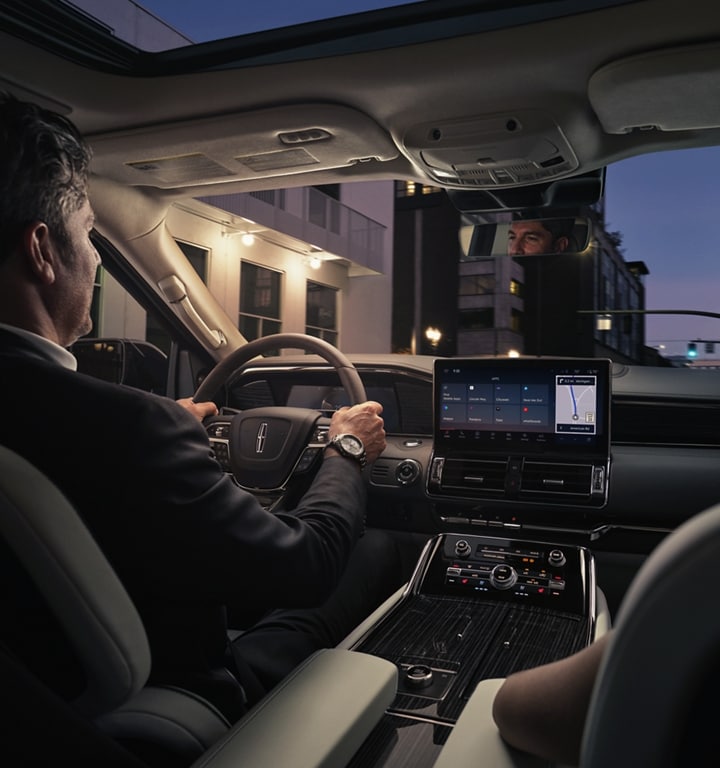 A man and woman are relaxing inside a 2024 Lincoln Navigator® SUV as they drive through a downtown street at night.