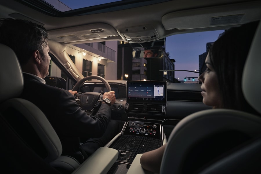 A man and woman are relaxing inside a 2024 Lincoln Navigator® SUV as they drive through a downtown street at night.