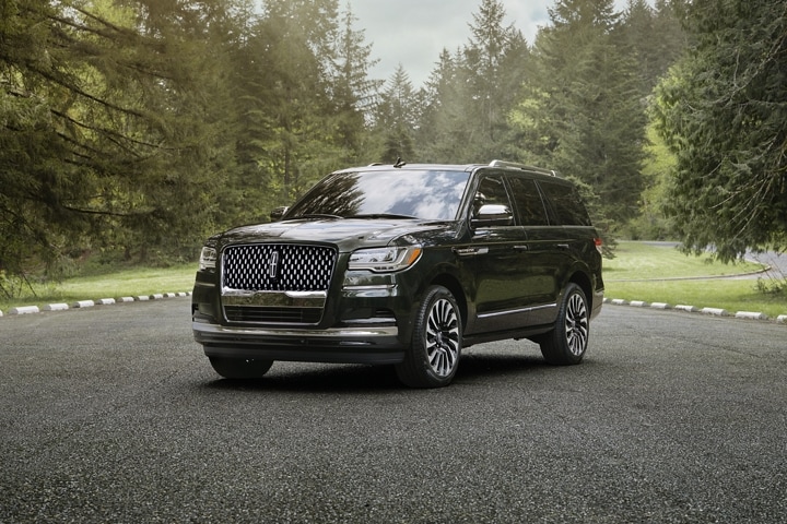 A 2024 Lincoln Black Label Navigator® SUV in Manhattan Green is parked on pavement near a scenic forest full of pine trees.