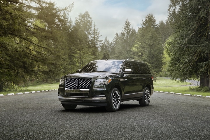 A 2024 Lincoln Black Label Navigator® SUV in Chroma Caviar is parked in a lot surrounded by lush forest and serene sunlight.