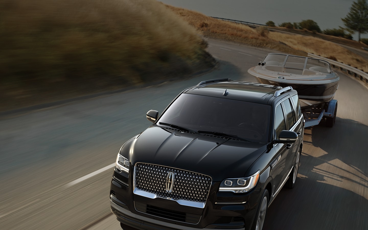 A 2024 Lincoln Navigator® SUV towing a boat is being driven along a curving road