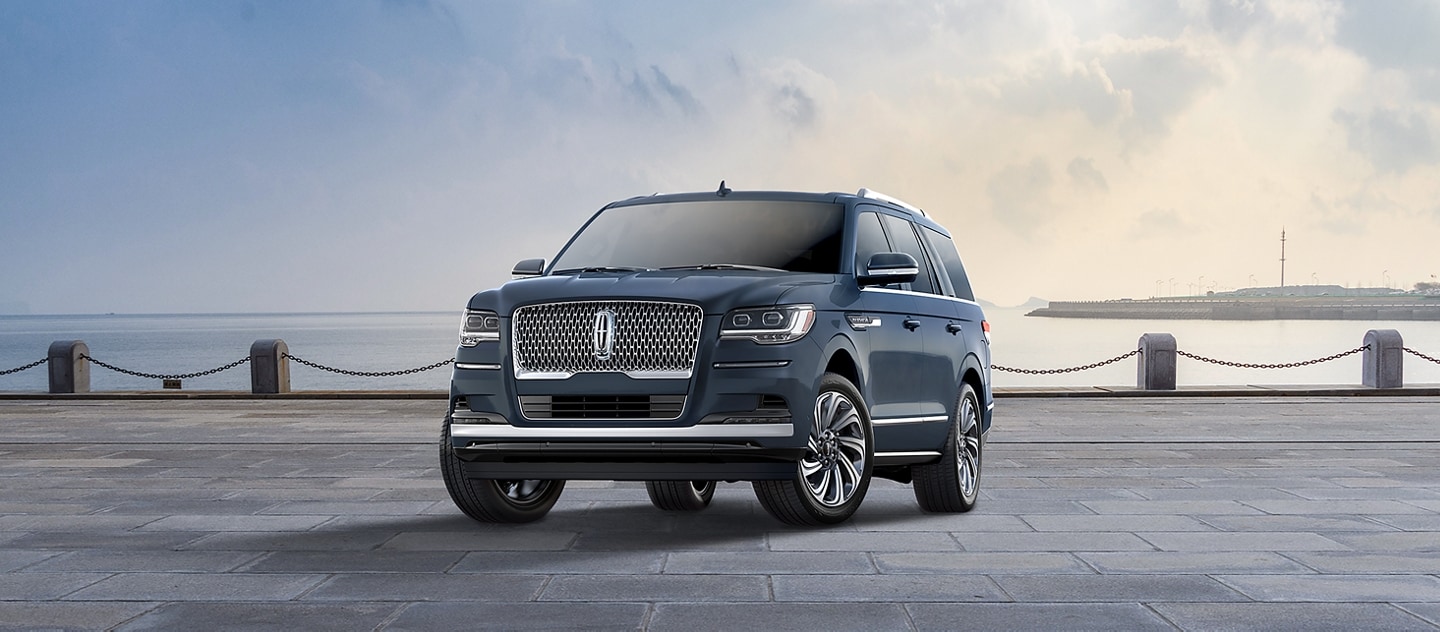 A 2024 Lincoln Navigator® Reserve model SUV in Flight Blue is parked near a bay beneath a cloudy sky.