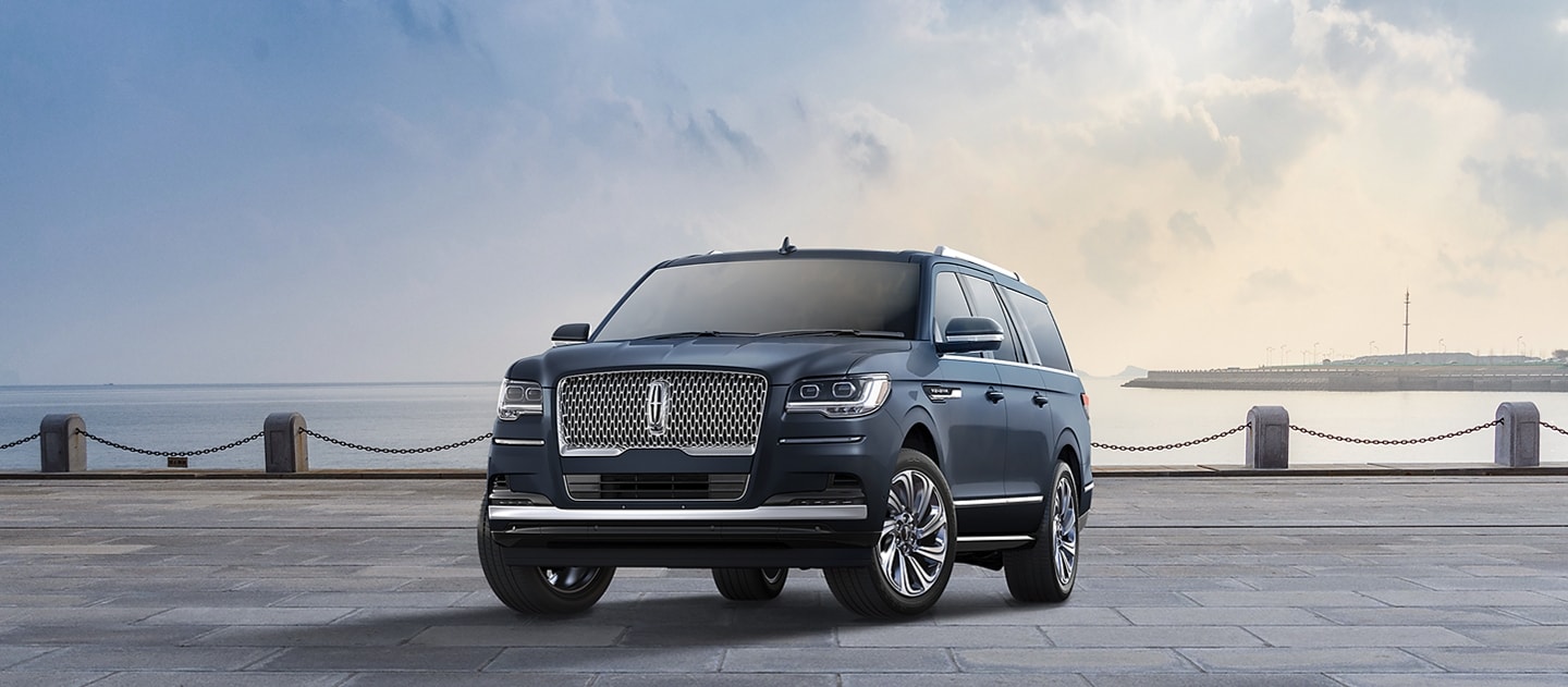 A 2024 Lincoln Navigator® Reserve L model SUV in Flight Blue is parked near a bay beneath a cloudy sky.