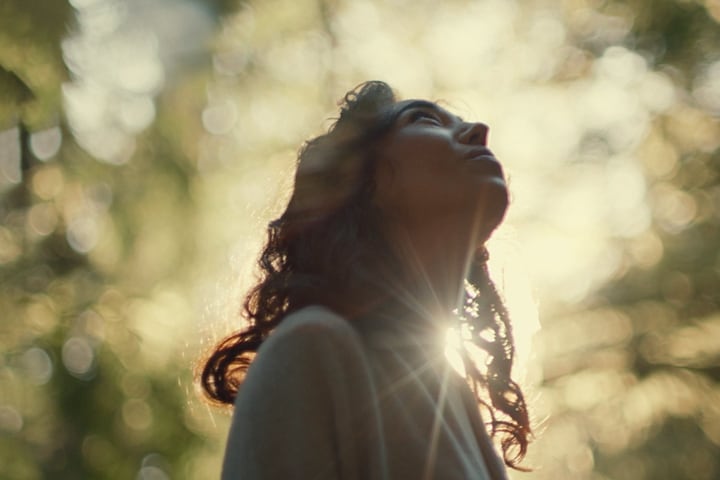 A woman is looking up as a sun flare and blurred trees behind her highlight her silhouette.