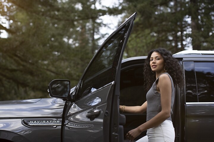 A woman is entering the driver's door of a 2024 Lincoln Black Label Navigator® SUV parked near a lush forest.